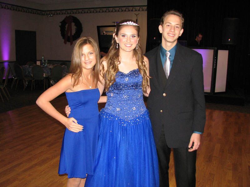 Three people posing for a photo. A woman in a blue gown and tiara stands between two people, all in formal attire.