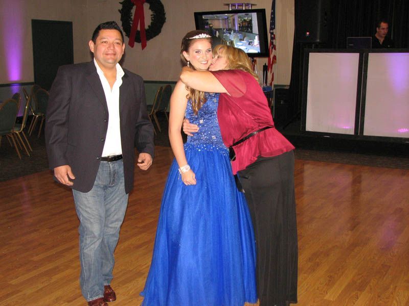 Man, teen in blue dress, and woman embrace in a room with a DJ booth, likely a dance or celebration.