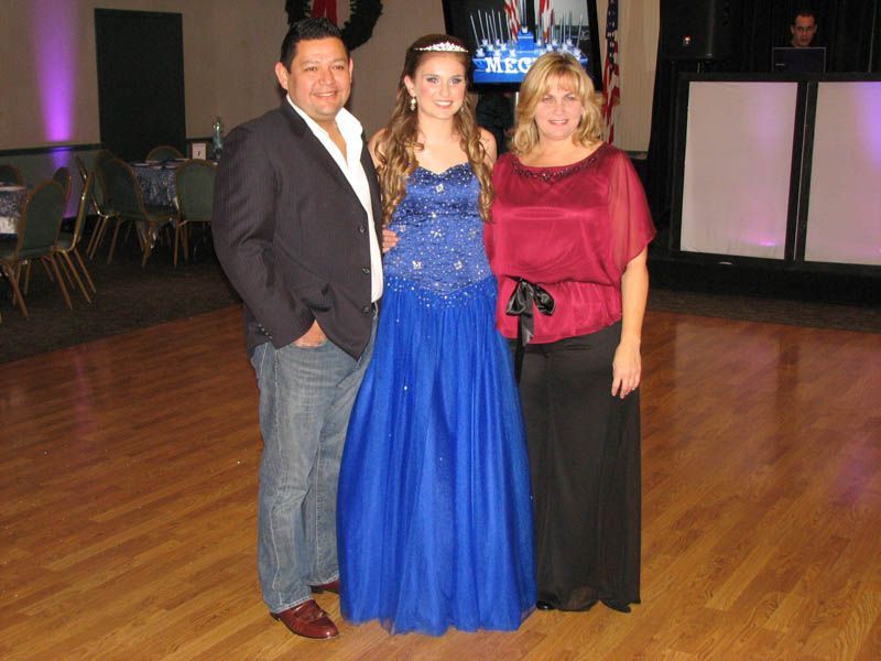 Family poses together in a room with a dance floor. Daughter in blue gown, parents smiling.