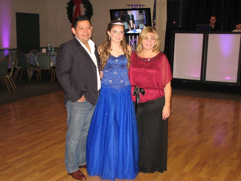 A family of three poses in a ballroom: a man in a blazer, a woman in red, and a girl in a blue dress.