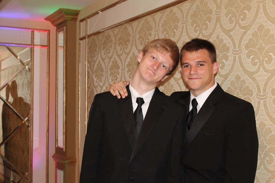 Two men in black suits and ties pose, one with arm around the other's shoulder, smiling against a patterned wall.