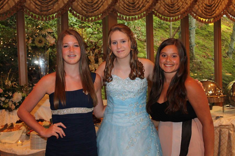 Three young people posing together at an event, one in a blue gown, others in party attire, near buffet tables.