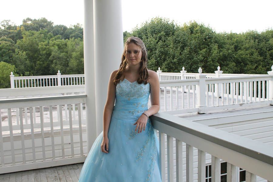 Woman in light blue formal dress on a white porch with trees in the background.
