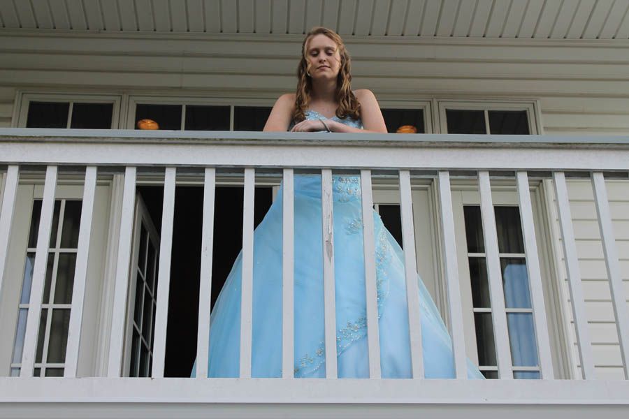 Woman in blue gown on a balcony, looking down with a smile. Balcony is white, house is light-colored.
