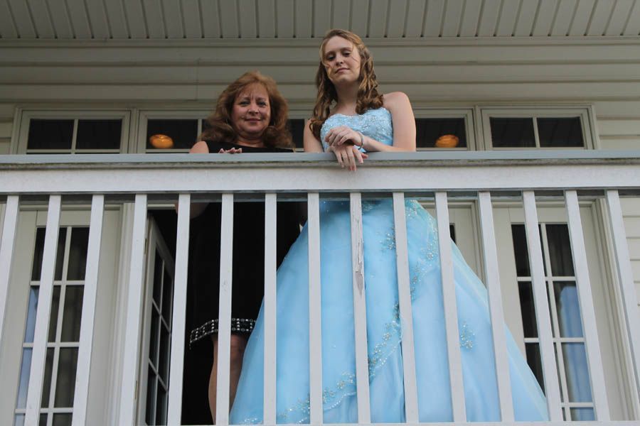Woman and teen in a light blue formal dress stand on a porch, hands on railing.