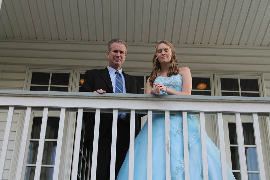 Man in suit and woman in blue gown on a porch, smiling.
