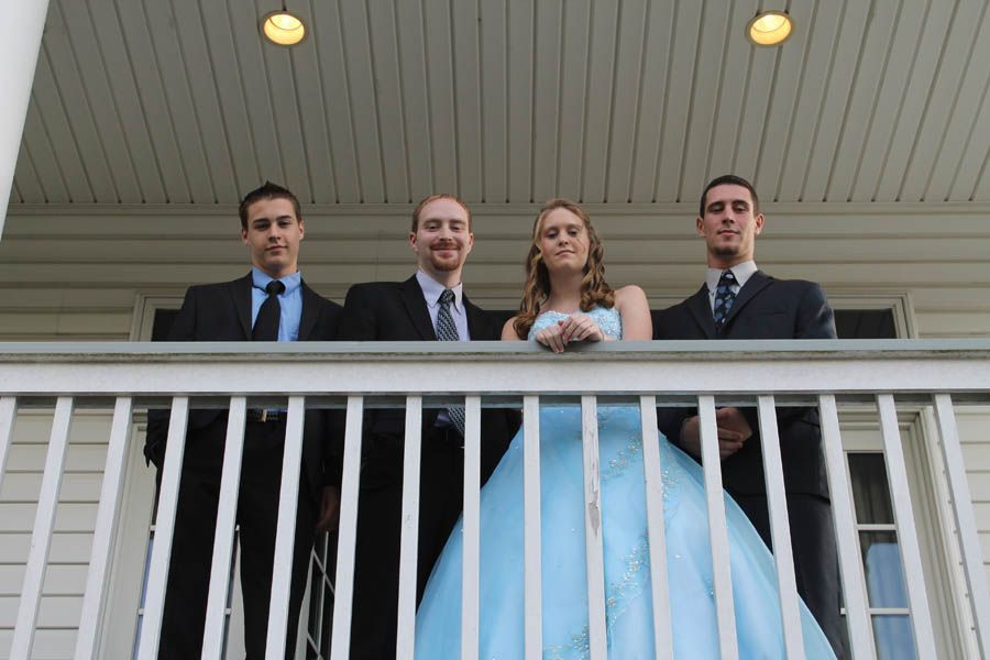 Four people on a porch. One woman in a blue gown, three men in suits. All are smiling.