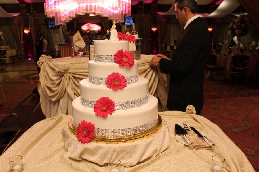 Wedding cake with pink flowers, silver trim, and a man in a suit cutting it at a reception.