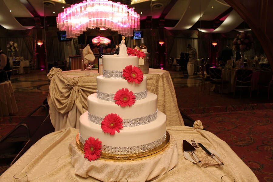 Four-tiered white wedding cake decorated with silver bands and pink flowers. Set on a golden tablecloth in a decorated hall.