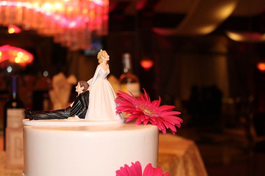 Wedding cake with bride and groom topper and pink flowers, in a dimly lit reception hall.