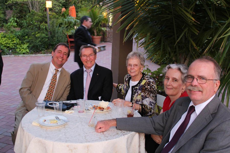Five people at a round table outside, smiling. Man in foreground points. Others are wearing suits and jackets.