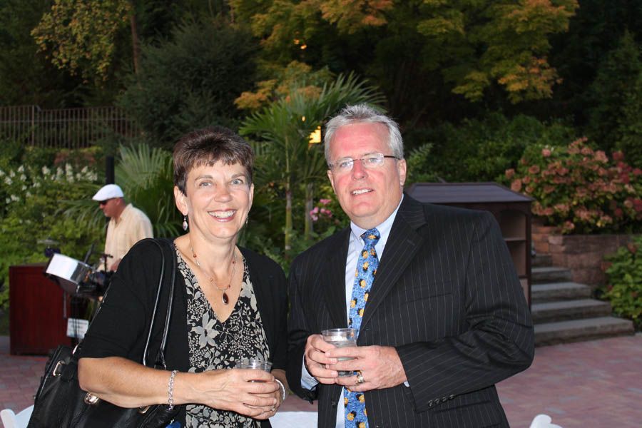 A couple smiles, holding drinks, outdoors near foliage. Man in suit, woman in black top.