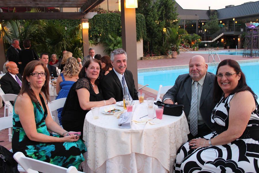 Group of people seated at a round table near a pool. Drinks, food, and smiles abound. Evening setting.