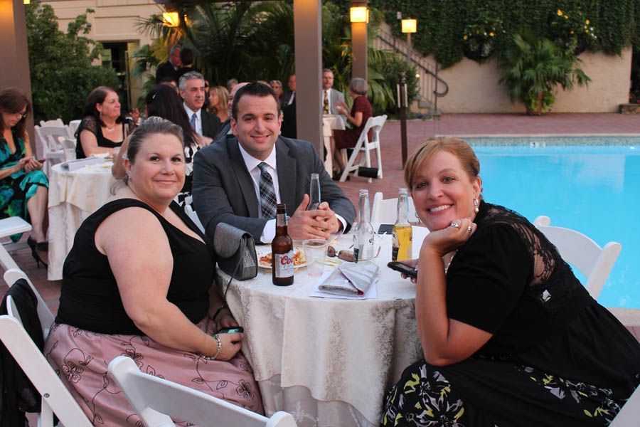 Three people at a table, smiling, near a pool. Man in suit, two women in dresses, outdoor setting.