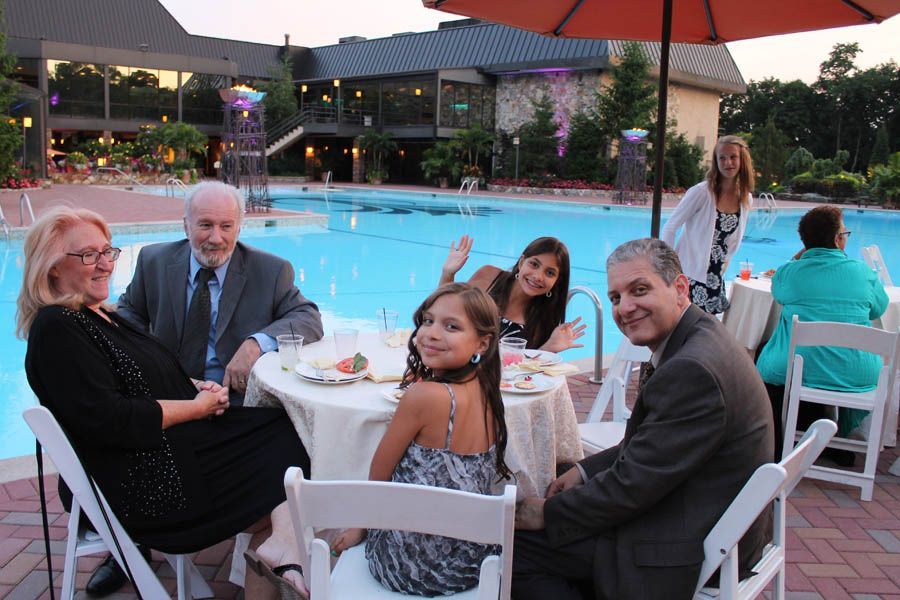 People seated at a table by a pool, one child waves. Building and trees in the background.
