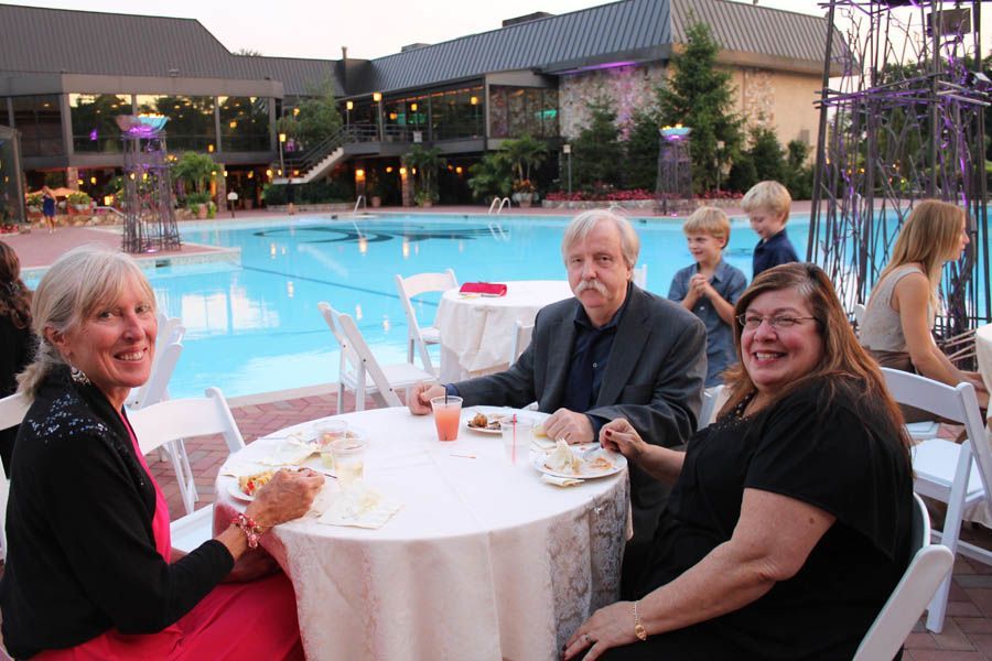 Three people seated at a round table by a pool, enjoying food and drinks. Building in background.