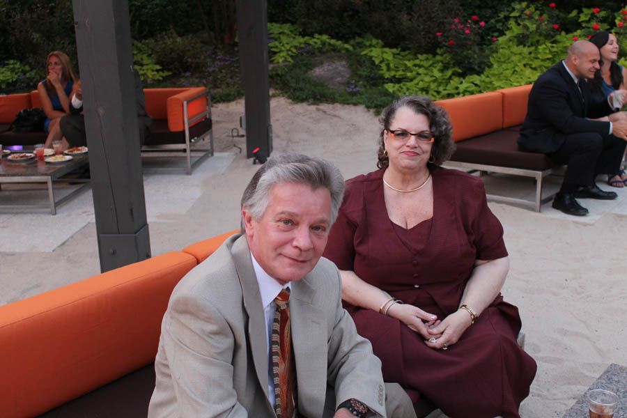 Man and woman seated on outdoor orange sofa, looking at the camera. Others in background.