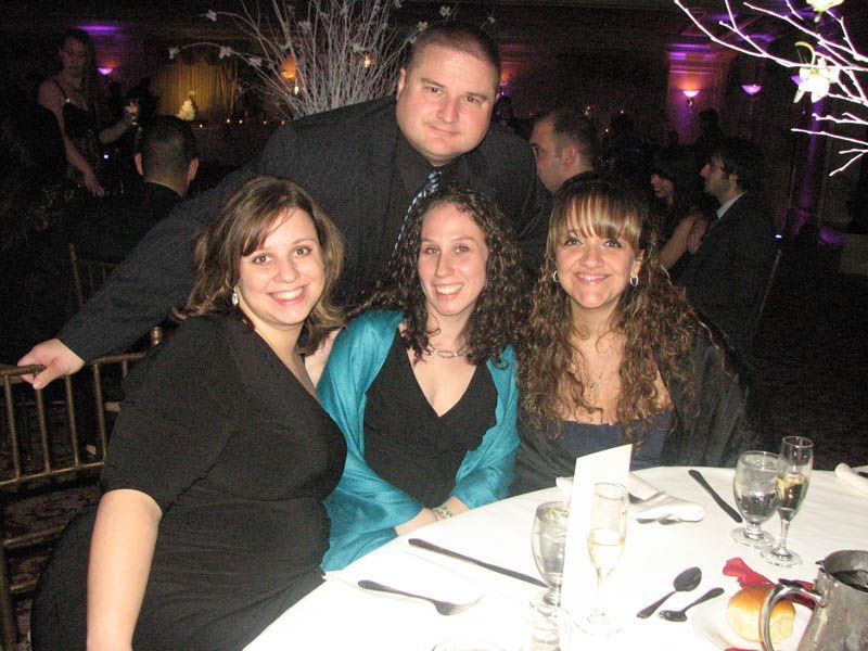 Four people at a table, smiling. Man standing behind them. Elegant party setting with decorations.