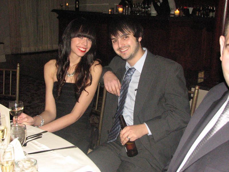 Woman in black dress and man in suit at a table. They are smiling at the camera, inside a dimly lit room.