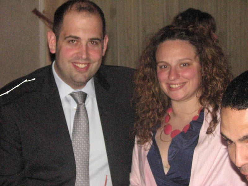 Man and woman smiling, indoors. Man in suit and tie. Woman wearing a pink shawl and necklace.