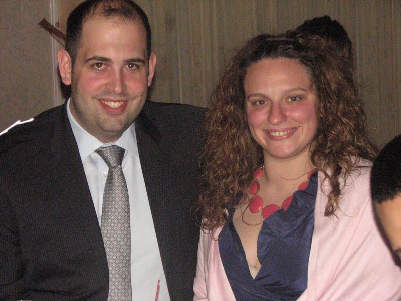 Man and woman smiling, indoors. Man in suit and tie. Woman in blue top, pink cardigan, and necklace.