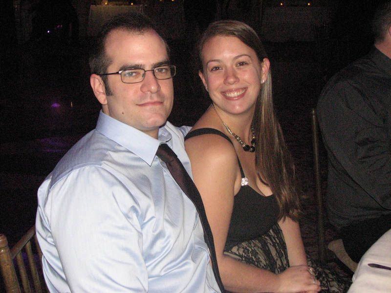 Man in glasses, light blue shirt, and tie, next to smiling woman in black dress, at an event.