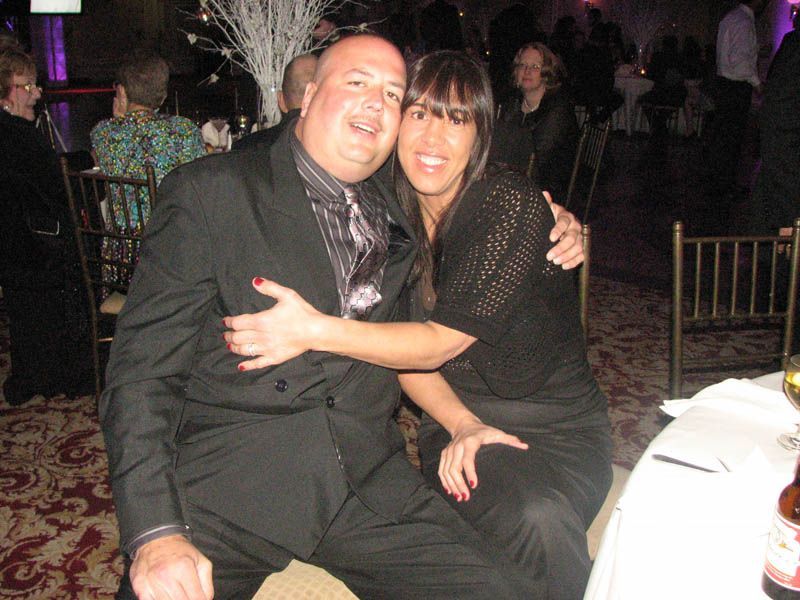 Man in suit embraces woman in black at an event. They both smile.