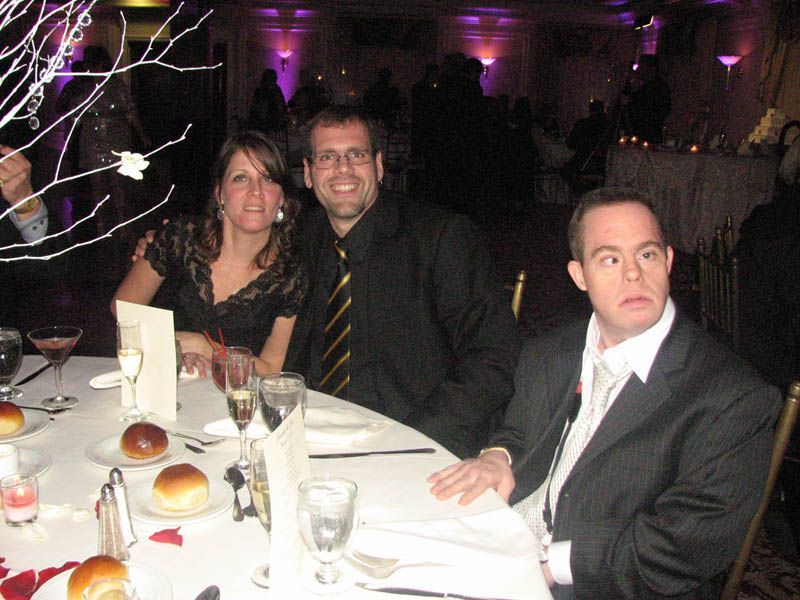 People seated at a formal dinner table: a couple smiling and a man looking to the side. Festive decorations in background.