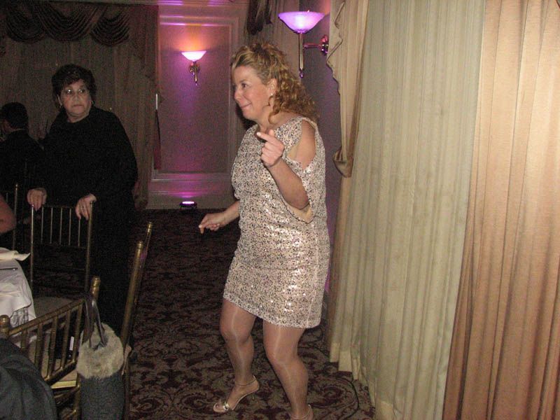 Woman in a sparkly dress dances near a curtain, another person watches. Dark carpet, indoor setting.