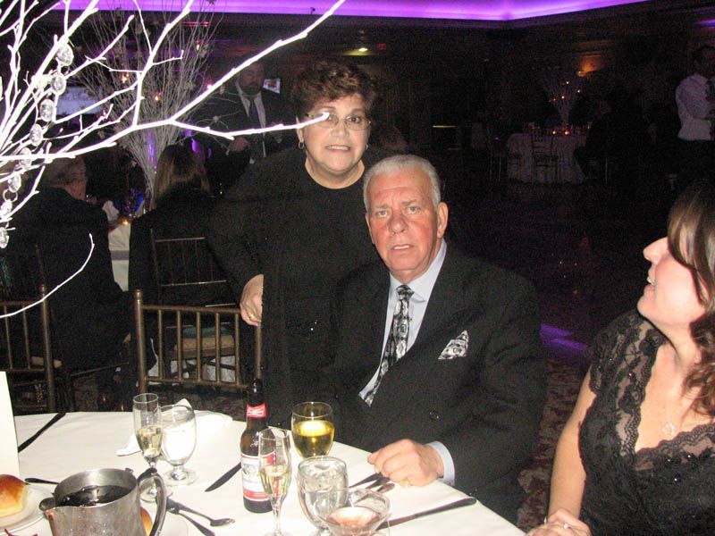 Two people at a table; woman standing behind man seated, both looking at camera. Festive setting with drinks and decorations.