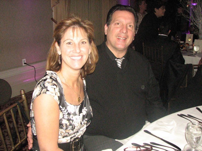 Woman in floral dress and man in black shirt at a table, smiling. Event setting.