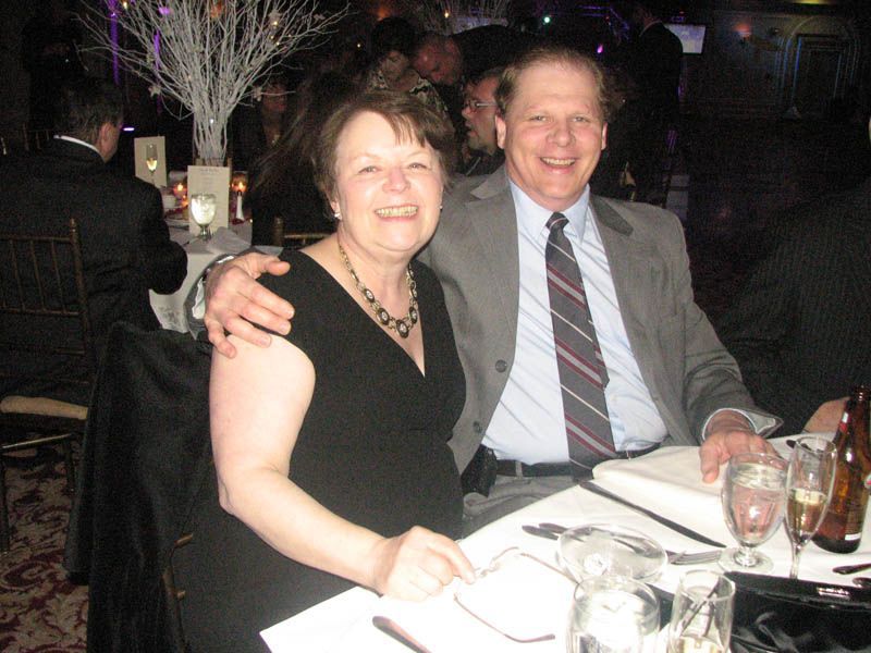 Couple smiling at a formal event; woman in black dress, man in suit, seated at a table.