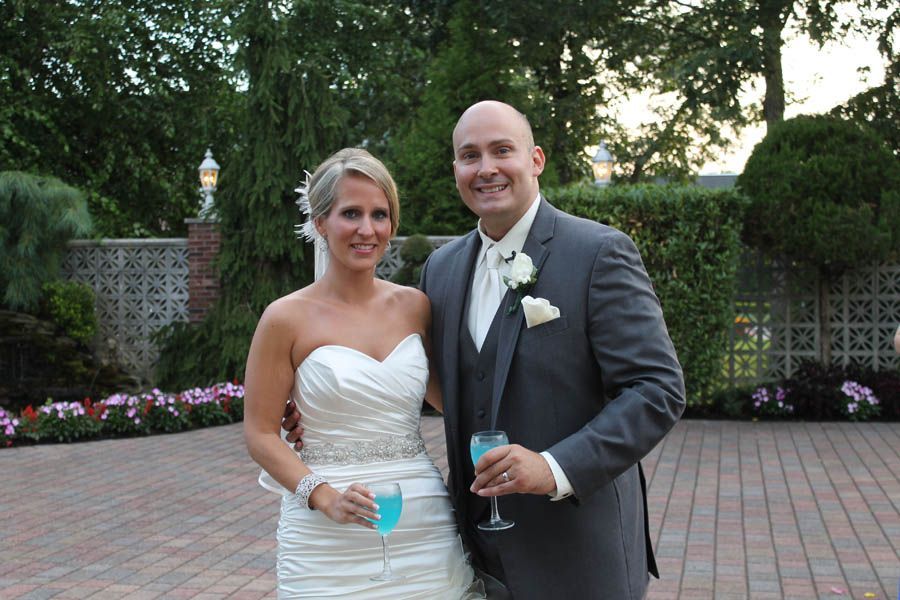 Bride and groom smiling, holding blue drinks, in formal attire on a brick patio with greenery.