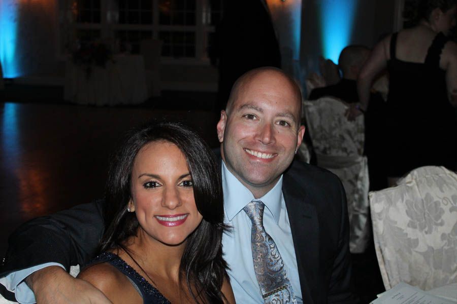 Smiling couple at an event; woman with dark hair, man in a suit with a patterned tie; event hall background.