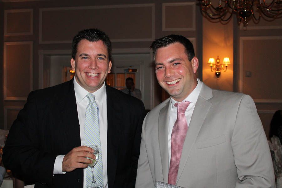 Two men in suits smiling, indoors. One holds a glass. Light pink and light blue ties.