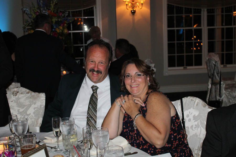 Couple at a formal event, smiling at a table set with glasses and floral arrangements.