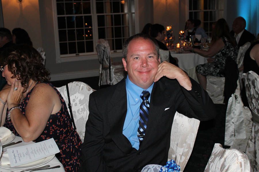 Man in a suit smiles, seated at a formal table. Others dine in the background, dimly lit.