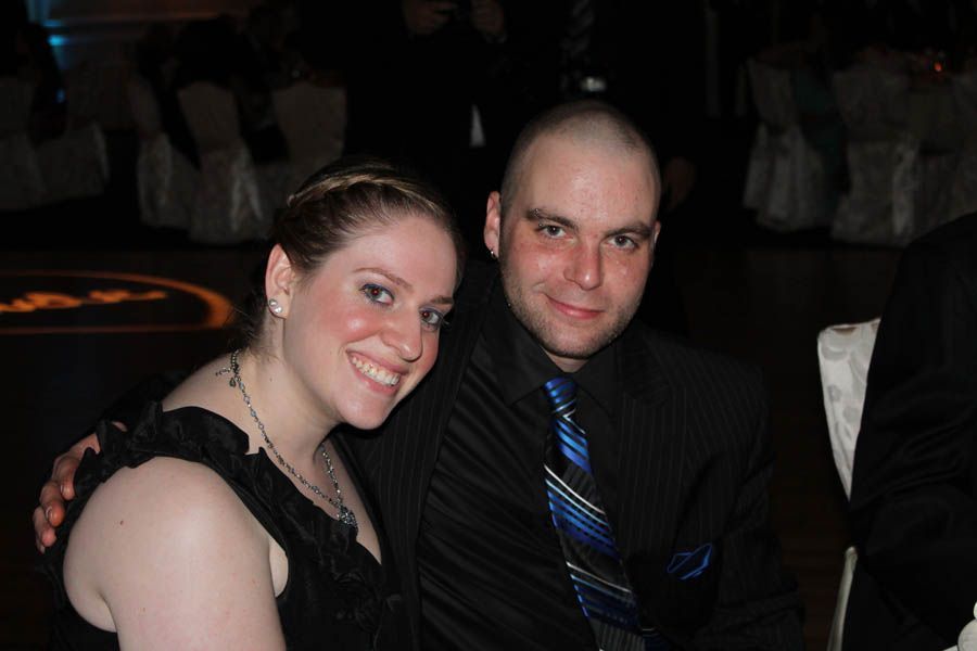 Woman and man smiling, seated close together at a formal event. Man in suit and tie. Woman in black dress.