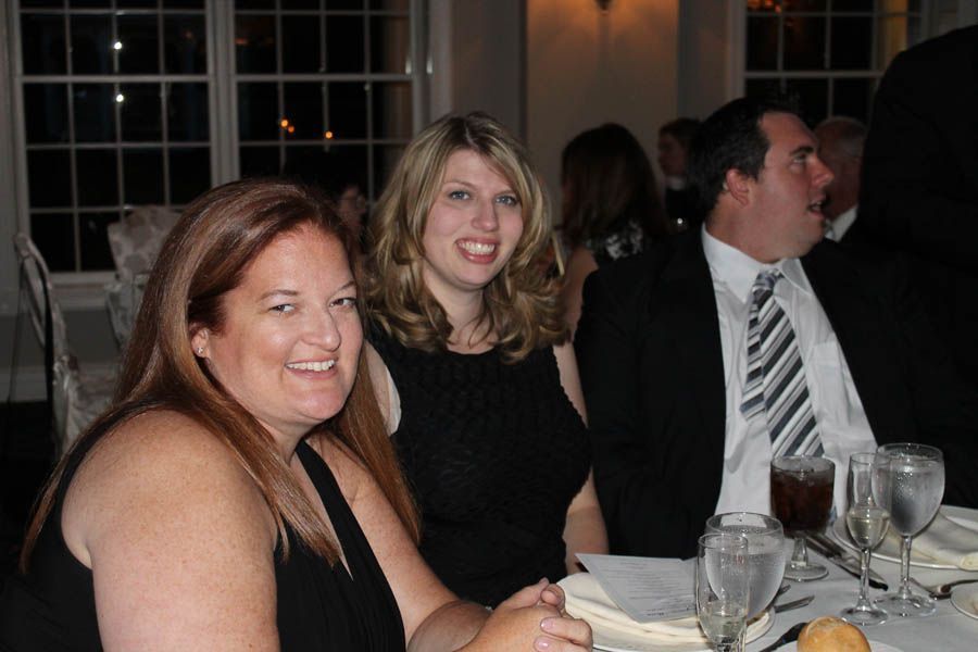 Three people at a formal dinner: two women and a man in a suit, seated at a table with drinks, smiling.