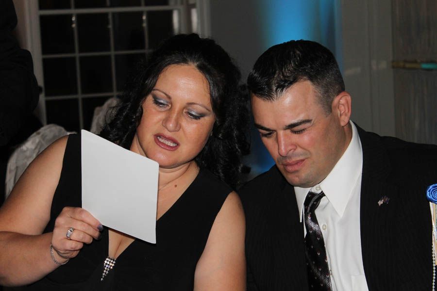 Woman in black dress reads a card to a man in a suit, both seated indoors.