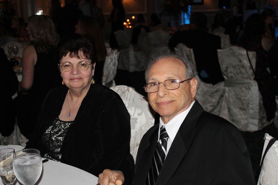Couple at a formal event, seated at a table. Man in suit, woman in black jacket, smiling.