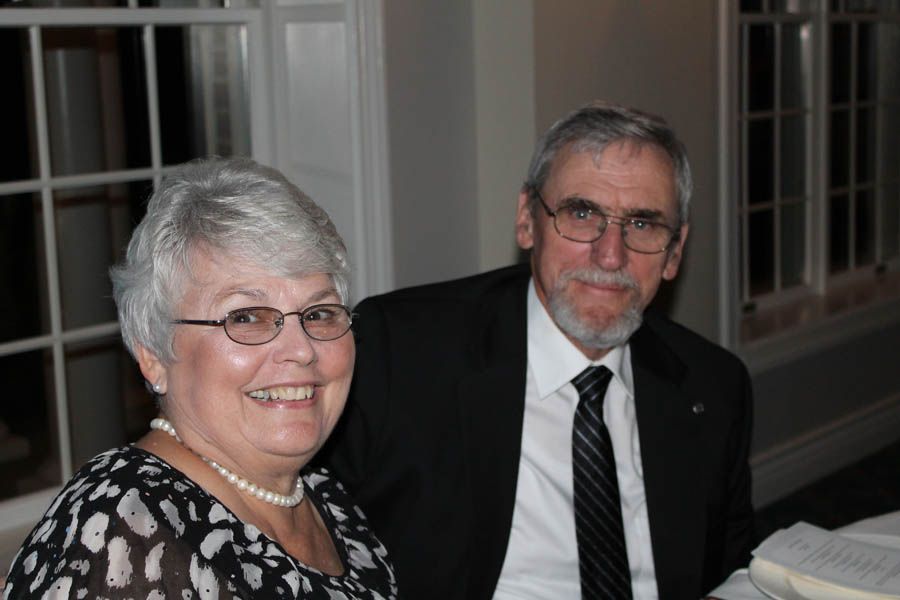 A smiling couple, both wearing glasses. Man in suit and tie. Woman in patterned blouse.