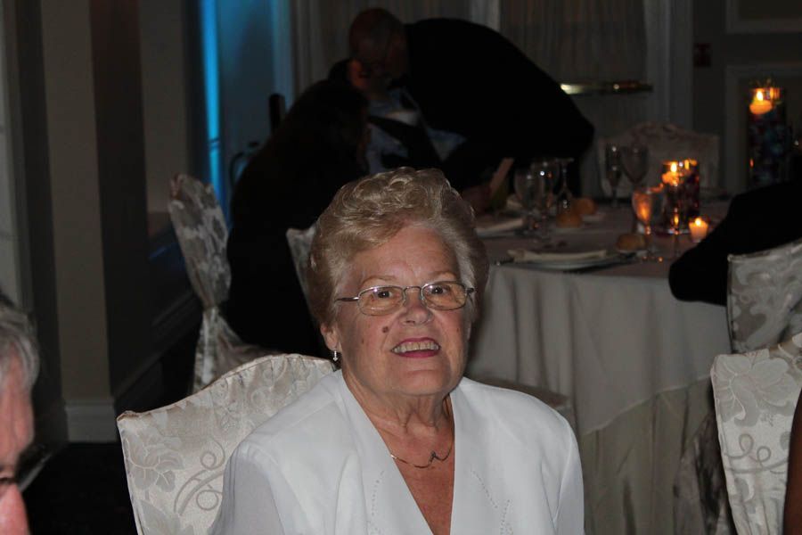 Woman with glasses at a table, wearing white. Others are at the table, some are blurred in the background.