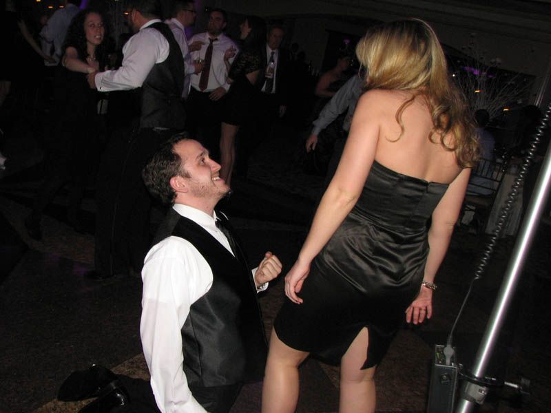 Man on one knee, looking up at woman in black dress on a dance floor.