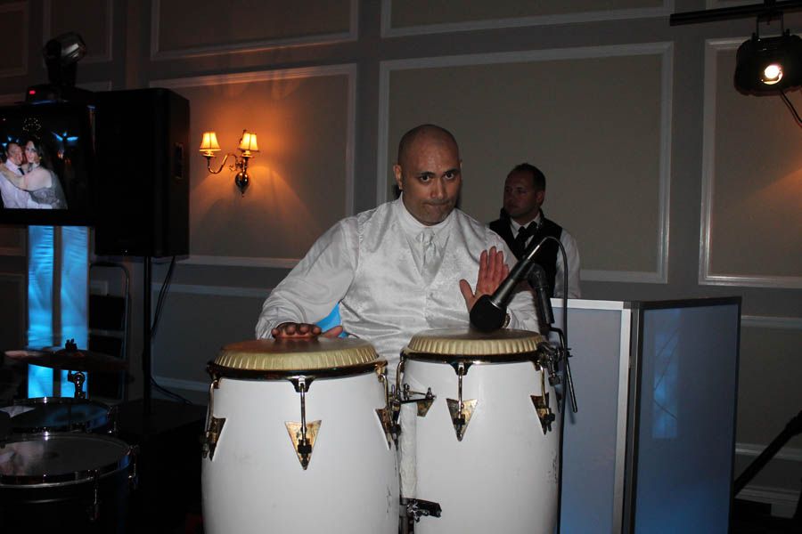 Man in white shirt playing conga drums at an event, with DJ setup in background.