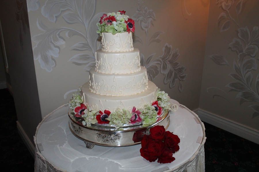 Five-tiered white wedding cake with floral decorations, on a silver stand, and red roses nearby.