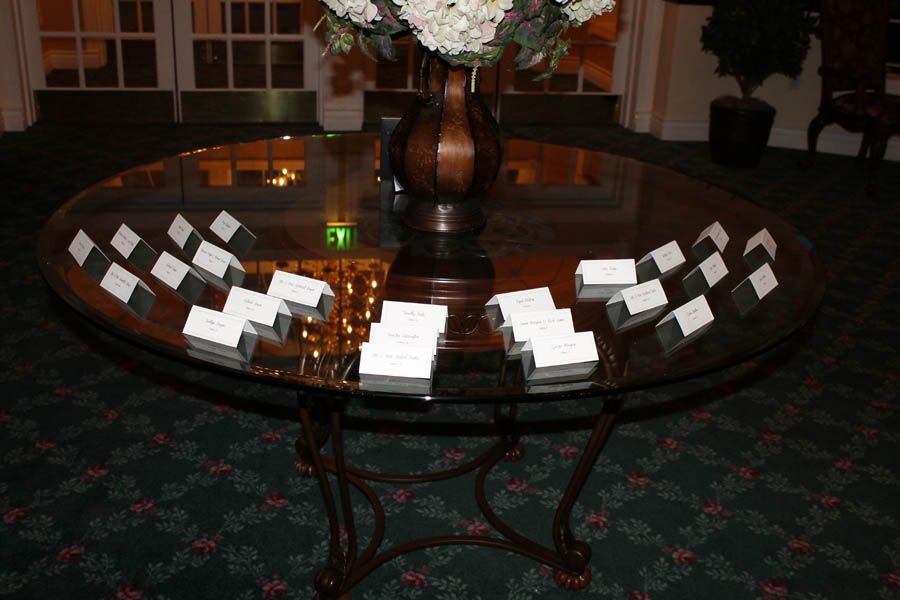 Round glass table with place cards arranged around a vase of white flowers, in a carpeted room.