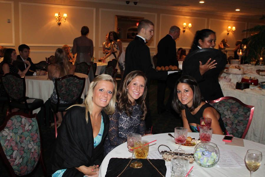 Three women smiling at a table in a dimly lit banquet hall. Others in formal wear are also present.