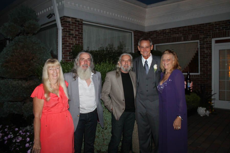 Five people pose outdoors: two women in red and purple dresses, three men with long hair and beards in suits and jackets.