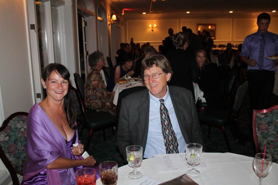 Woman in purple dress and man in suit at a formal dinner table, others seated in the background.